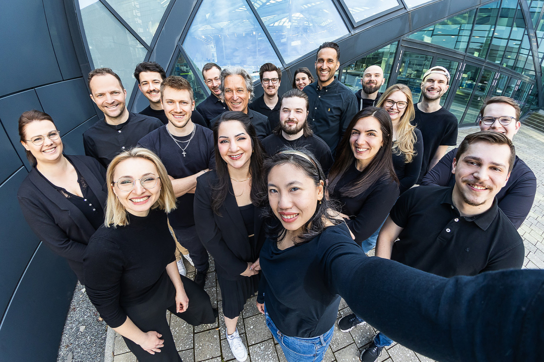 Selfie-Aufnahme des mindtwo-Teams im Au&szlig;enbereich vor einem modernen B&uuml;rogeb&auml;ude mit Glasfassade.