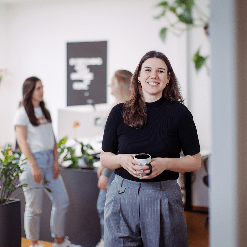 Eine Person steht lächelnd mit einer Kaffeetasse im Vordergrund, während im Hintergrund drei Kolleginnen miteinander sprechen.