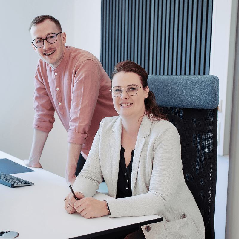 Zwei Personen arbeiten gemeinsam an einem Schreibtisch im Büro und blicken freundlich in die Kamera.