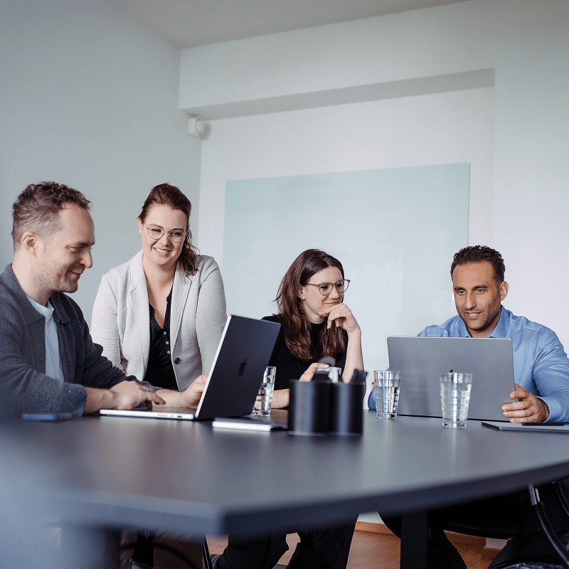 Vier Personen sitzen an einem Konferenztisch mit Laptops und besprechen gemeinsam ein Projekt in einer modernen Büroumgebung.